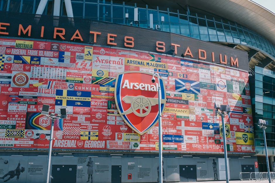 A large exterior view of the Emirates Stadium featuring a prominent red and white Arsenal logo shield at the center. The stadium facade is decorated with a collage of numerous Arsenal supporter banners, flags, and patches in various colors, displaying team names, symbols, and supporters' slogans. The building's modern architectural design includes extensive glass windows and metallic panels, with bright stadium lighting and security cameras mounted on poles visible in the foreground. The scene captures a vibrant, bustling atmosphere typical of a sports venue, with clear daylight illuminating the detailed signage and decorations.