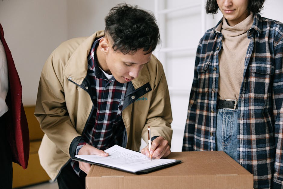 A person with medium skin tone, wearing a beige jacket over a plaid shirt, is seen bent over a cardboard box while using a pen to fill out a checklist on a clipboard. Next to them, another individual with light skin, dressed in a blue and beige plaid shirt and high-waisted jeans, stands observing. The setting appears to be an indoor space with neutral-colored walls, and natural lighting highlights the cleanliness and orderliness of the area. This scene depicts a detailed moment of a property inspection, aligned with surface cleaning, sanitisation, and maintenance tasks typical of end of tenancy cleaning procedures, as referenced on the page titled 'Drayton Park Highbury end of tenancy cleaning checklist' from Oven Cleaning Highbury.