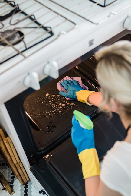 A person wearing blue and yellow rubber gloves is cleaning the interior of a black oven, wiping the oven's glass door with a pink cloth. The oven is part of a white gas stove with visible burners and control knobs on top. The kitchen floor features black and white patterned tiles. The scene highlights surface cleaning and sanitisation processes performed by Oven Cleaning Highbury, emphasizing cleanliness and maintenance in a domestic kitchen environment.