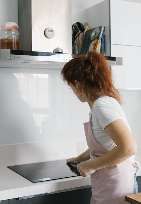 A woman with red hair, dressed in a white t-shirt and light pink apron, is cleaning a black glass induction cooktop in a modern kitchen. The kitchen features white glossy cabinets and a white countertop, with a stainless steel range hood above the stove. On the left, there are glass jars with ingredients on the counter, and a digital thermometer on the wall. The lighting is natural and bright, highlighting the clean and tidy surface, with no visible dirt or stains. This scene exemplifies domestic surface cleaning and maintenance, consistent with the services provided by Oven Cleaning Highbury, emphasizing hygiene and pristine kitchen surfaces.
