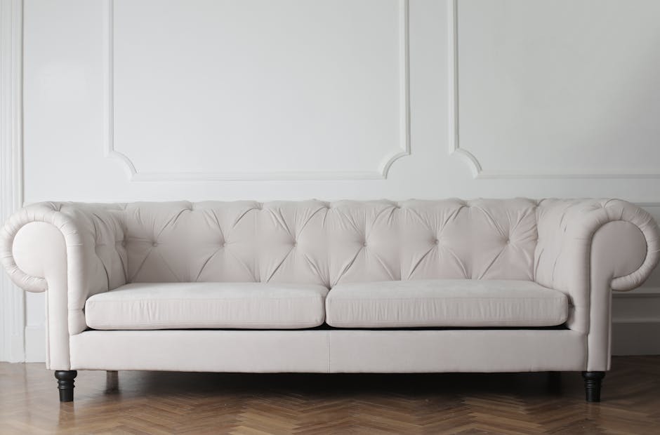 A white, upholstered sofa with button-tufted cushions and rolled arms positioned against a white decorative wall panel in a room with wooden flooring. The sofa appears clean and well-maintained, with no visible stains or dust. The background features a simple, elegant wall design, and the lighting is soft, highlighting the smooth fabric and tidy appearance. This scene exemplifies the type of surface cleaning and domestic maintenance encouraged by Oven Cleaning Highbury, emphasizing hygiene and furniture care in a home setting.
