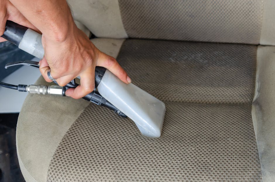 A person using a handheld steam cleaner to deep clean a beige fabric-upholstered armchair in a residential living room. The steam cleaner, featuring a gray nozzle and black handle, is pressed onto the textured fabric surface, which shows visible dirt and stains. The room is well-lit, with natural light highlighting the sofa's smooth armrest and cushion, which has a subtle woven pattern. The background includes part of the sofa's wooden frame and a soft fabric cushion. The image demonstrates surface cleaning and stain removal as part of routine domestic cleaning, with the process emphasizing hygiene and maintenance, consistent with services offered by Oven Cleaning Highbury.