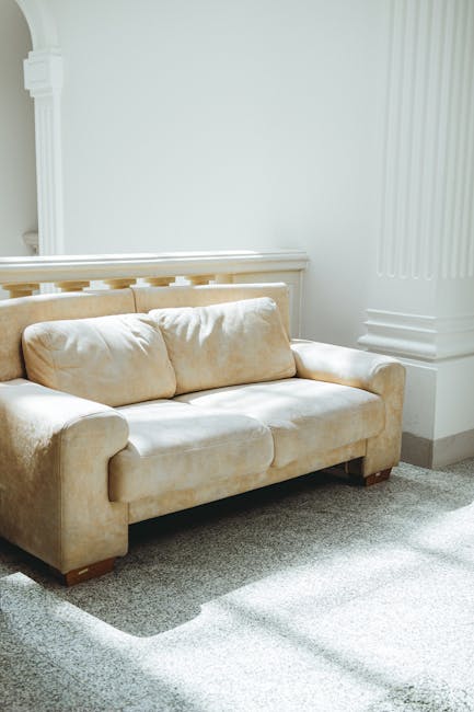 A white, upholstered sofa with button-tufted cushions and rolled arms positioned against a white decorative wall panel in a room with wooden flooring. The sofa appears clean and well-maintained, with no visible stains or dust. The background features a simple, elegant wall design, and the lighting is soft, highlighting the smooth fabric and tidy appearance. This scene exemplifies the type of surface cleaning and domestic maintenance encouraged by Oven Cleaning Highbury, emphasizing hygiene and furniture care in a home setting.