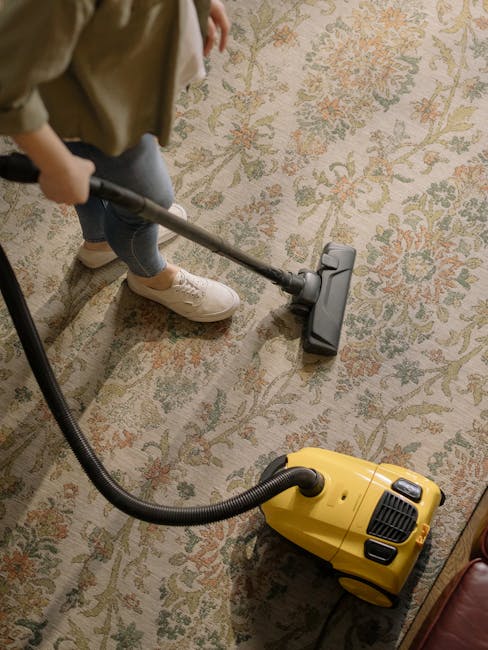 A professional cleaning technician dressed in full white protective coveralls, face mask, and blue gloves performs deep cleaning on a carpet inside a domestic living room. The technician uses a specia