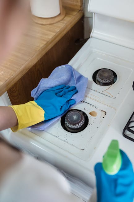 A person wearing yellow and blue rubber cleaning gloves is using a blue cloth for surface cleaning on a white electric stovetop with two burners. The stovetop shows some visible stains and dirt around the burners, indicating a need for deep cleaning. Leaning against a wooden kitchen counter, which holds a white container, the scene is well-lit, emphasizing the cleanliness process. The photograph showcases routine domestic cleaning, with a focus on sanitising and maintaining the kitchen surfaces. The image is associated with Oven Cleaning Highbury, highlighting the importance of thorough surface cleaning and hygiene in kitchen maintenance.