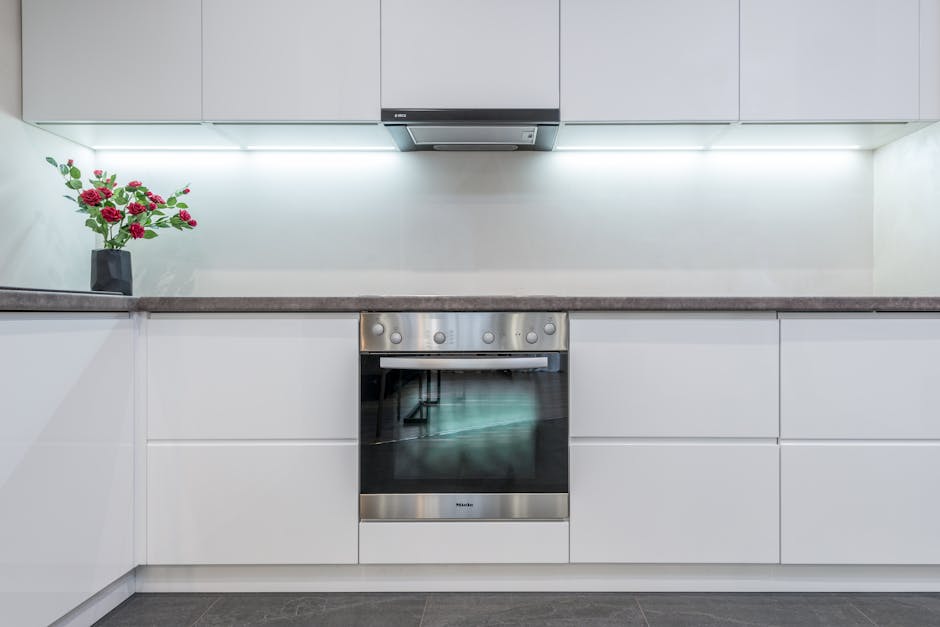 A modern kitchen with white flat-front cabinets and a dark countertop, featuring a stainless steel oven integrated into the cabinetry beneath the counter. The wall above the counter is plain and painted white, accented by under-cabinet lighting that illuminates the workspace. On the left side, there is a black vase holding a bouquet of pink roses with green foliage, positioned on the counter. The kitchen appears clean and well-maintained, with no visible dirt or stains on surfaces, exemplifying surface cleaning and hygiene. The space is well-lit with natural or artificial light, highlighting the sleek, minimalist design typical of deep cleaning and sanitisation processes, as promoted by Oven Cleaning Highbury.
