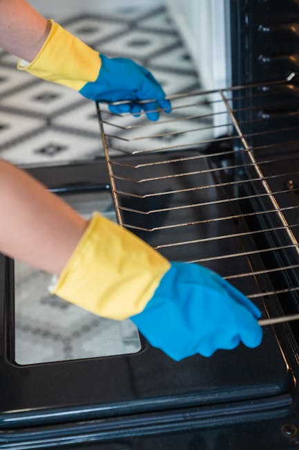 Close-up of a person wearing yellow and blue rubber gloves cleaning the interior of a black oven with a wire rack, which appears to be free of grease and stains. The oven's ceramic or enamel surface is visible around the door frame, and the scene is lit with ambient light highlighting the clean, shiny glass door. The background hints at a tiled kitchen floor with a geometric black and white pattern, emphasizing the household setting. This image illustrates surface cleaning and deep sanitation processes undertaken by Oven Cleaning Highbury as part of their domestic cleaning services, aligning with the focus of the 'Same day oven cleaning Highbury and Islington booking guide' page.