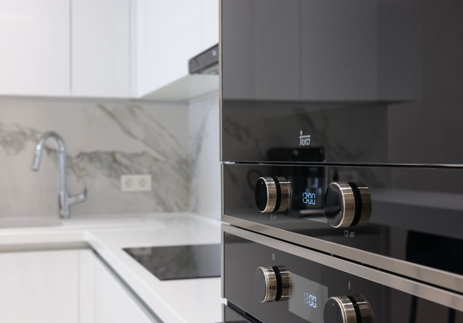 Close-up view of a modern kitchen oven with a sleek black stainless steel finish and illuminated digital controls, situated within a clean, minimalist kitchen setting. The oven's surface appears spotless and well-maintained, with reflective surfaces contrasting against the white marble backsplash and countertops. To the left, a white sink with a silver faucet is visible, along with ceramic tiles and an electrical outlet on the backsplash. The overall scene highlights a well-organized, hygienic environment suitable for surface cleaning and sanitisation, with natural light enhancing the pristine condition. Oven Cleaning Highbury provides expert domestic cleaning services to ensure appliances like this oven are deep cleaned and optimised for hygiene.